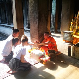 May include: A Buddhist monk in orange robes sits on the floor of a temple, accepting offerings from a family. The family is kneeling and holding a string of beads. The monk is holding a bowl and a small box. There is a sign that says "OBJECT DONATION" in the background.