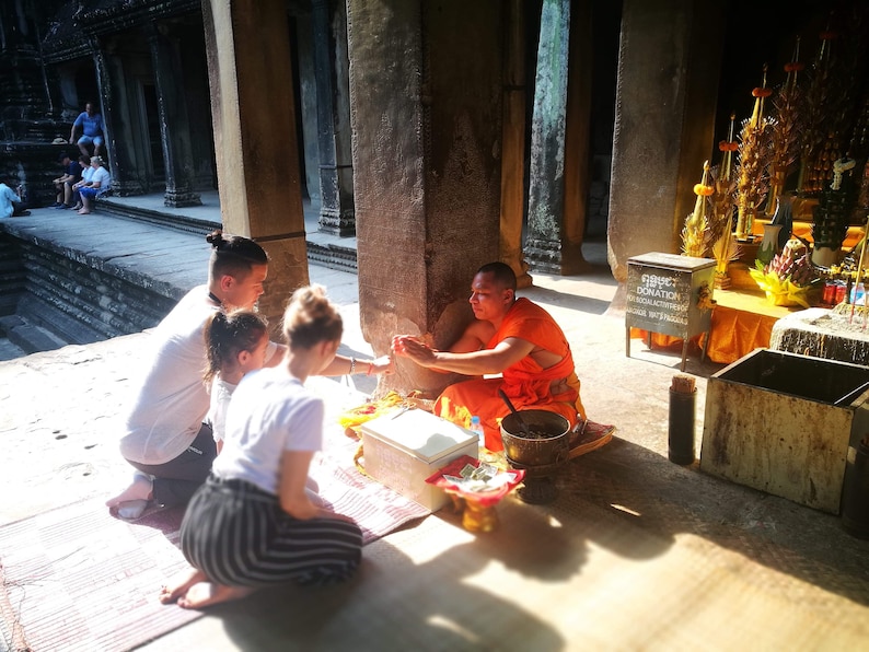 May include: A Buddhist monk in orange robes sits on the floor of a temple, accepting offerings from a family. The family is kneeling and holding a string of beads. The monk is holding a bowl and a small box. There is a sign that says "OBJECT DONATION" in the background.