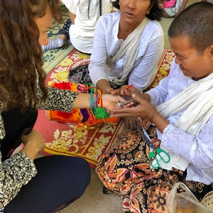 May include: A woman ties colorful string bracelets on the wrist of another woman. The woman tying the bracelets is wearing a white shirt and a long white scarf. The woman receiving the bracelets is wearing a white shirt and a long black and orange skirt.