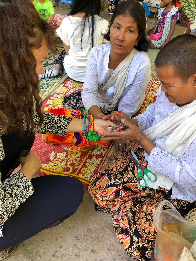 May include: A woman ties colorful string bracelets on the wrist of another woman. The woman tying the bracelets is wearing a white shirt and a long white scarf. The woman receiving the bracelets is wearing a white shirt and a long black and orange skirt.