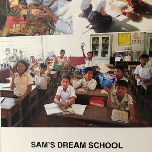 May include: A group of children in a classroom setting, some holding books and others looking at the camera. The classroom is brightly lit and has a water cooler and a sign that reads "Sam's Dream School, Siem Reap, Cambodia".