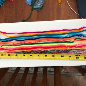 May include: A close-up of a white surface with a row of colorful friendship bracelets laid out. A yellow measuring tape is placed below the bracelets, showing measurements from 1 to 15 inches.