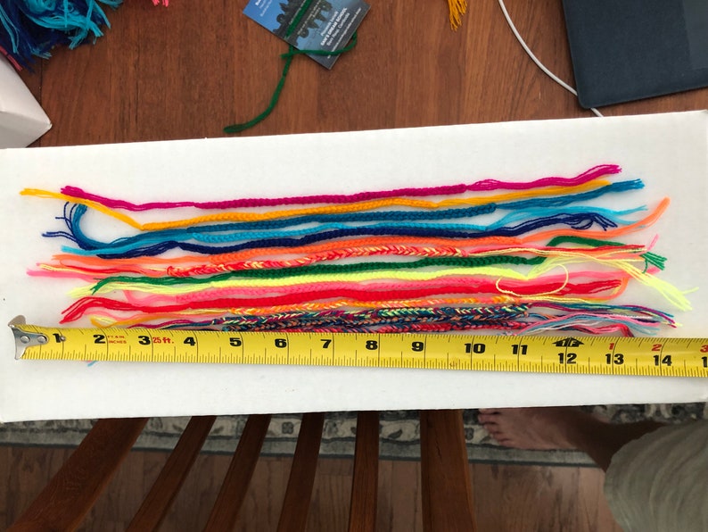 May include: A close-up of a white surface with a row of colorful friendship bracelets laid out. A yellow measuring tape is placed below the bracelets, showing measurements from 1 to 15 inches.
