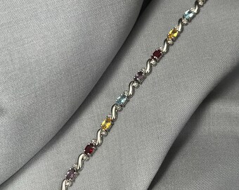 Può includere: Un bracciale in argento con una serie di pietre preziose ovali di vari colori, tra cui rosso, viola, giallo e blu. Le pietre sono incastonate in un motivo ondulato con piccole pietre chiare. Il bracciale è esposto su un tessuto grigio.