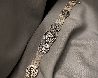 May include: A silver bracelet with multiple chains and decorative round medallions. The medallions have a delicate, intricate design.