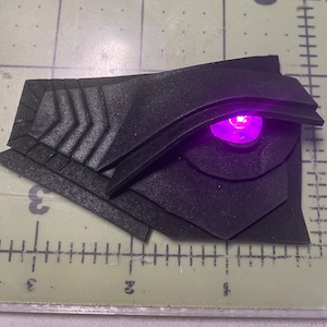 May include: A black 3D printed eye with a glowing purple LED light. The eye is detailed with ridges and a textured surface.