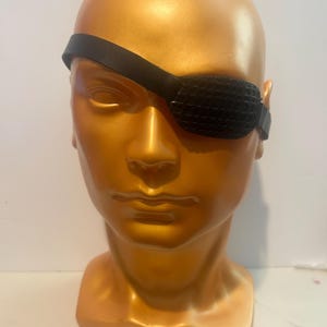 May include: A black eye patch with a textured grid pattern on the front, secured by a smooth black strap. The eye patch is displayed on a metallic gold-colored mannequin head that features defined facial contours, including eyes, nose, and lips.