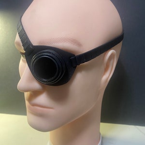 May include: A black eye patch featuring a circular, raised lens-like component over the eye area. The patch is secured by a textured, adjustable black strap that wraps around the head, providing a structured fit over the eye socket.