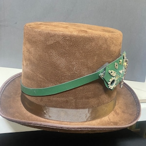 Può includere: Un cappello a cilindro in pelle scamosciata marrone con una fascia in pelle verde e un ornamento in metallo di ispirazione steampunk.
