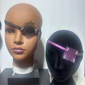 Puede incluir: Dos parches decorativos para los ojos en cabezas de maniquí. Uno es negro con un diseño de rejilla y detalles plateados, el otro es morado con un diseño de rejilla y detalles plateados. Los parches para los ojos están sujetos a la cabeza con una correa.