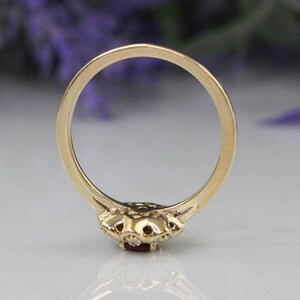May include: A gold ring with a red gemstone surrounded by smaller white gemstones. The ring is shown from the back, revealing the smooth band.