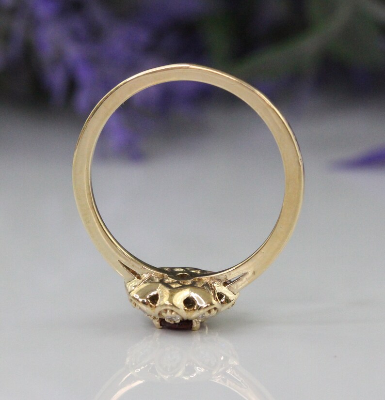 May include: A gold ring with a red gemstone surrounded by smaller white gemstones. The ring is shown from the back, revealing the smooth band.