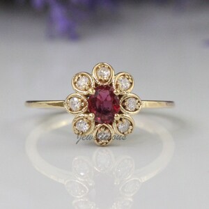 May include: A gold ring with a red oval gemstone surrounded by a halo of smaller white gemstones. The ring is set in a flower-shaped design.