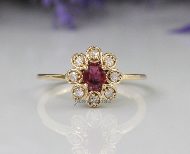 May include: A gold ring with a red oval gemstone surrounded by a halo of smaller white gemstones. The ring is set in a flower-shaped design.