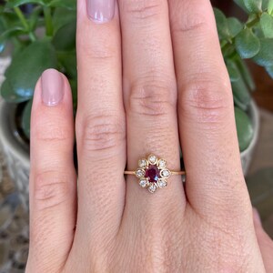 May include: A gold ring with a red gemstone surrounded by smaller white gemstones, set in a flower-shaped design.