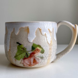 May include: A ceramic mug with a unique design. The mug features a white, dripped glaze around the rim and a floral illustration. The handle is curved and matches the mug's color. The mug is a beige color with a crackle finish.