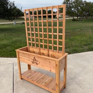 May include: A wooden planter box with a trellis, perfect for a garden. The planter is made of light brown wood and features a lower shelf. The trellis is a grid pattern. The planter has the word "Arness" engraved on the front.