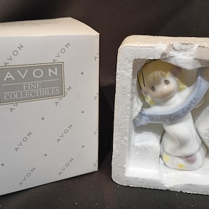 May include: A porcelain figurine of a child with blonde hair wearing a white robe, holding a light blue banner with the text Peace On Earth, packaged in a white styrofoam insert next to a white box labeled Avon Fine Collectibles.
