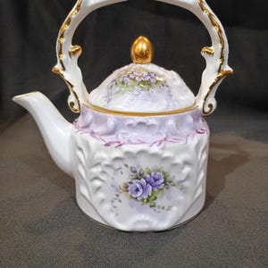 May include: A white porcelain teapot featuring a raised, embossed texture, painted with purple rose floral motifs on the body and lid. The teapot includes a gold-trimmed handle, a gold-finished finial on the lid, and a gold-rimmed spout opening.