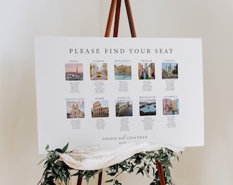 May include: A white seating chart with a black and white title "PLEASE FIND YOUR SEAT". The chart lists cities around the world with a small photo of a landmark in each city. The chart is on an easel with greenery and a white fabric draped over it.