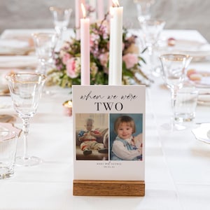 May include: A white table card with the text "when we were two" and two photos of children. The card is on a wooden stand and is on a white table with a floral centerpiece.