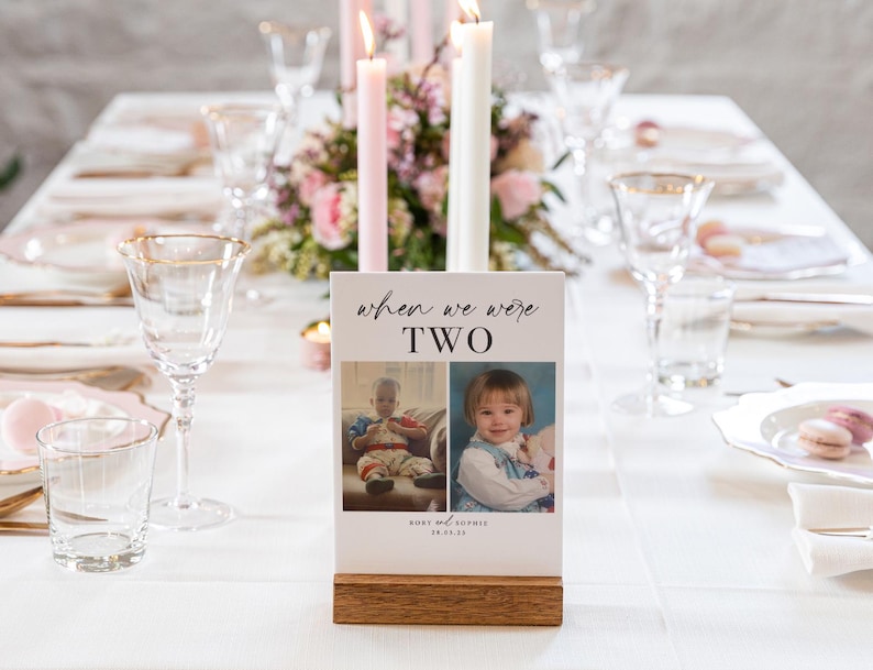 May include: A white table card with the text "when we were two" and two photos of children. The card is on a wooden stand and is on a white table with a floral centerpiece.