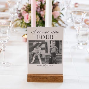 May include: A white card with black text that reads "when we were FOUR" with a black and white photo of two children. The card is on a wooden stand and is on a white tablecloth.