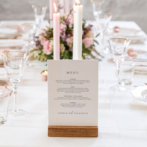May include: A white table setting with a menu card on a wooden stand. The menu card has the text "MENU" at the top and the names "Sophie & Jonathan" at the bottom. The table is set with white plates, glasses, and pink flowers.