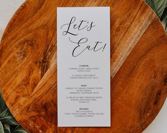 May include: A white card with black text on a wooden surface. The card has the words "Let's Eat!" at the top and lists a menu with starter, main, and dessert options. The menu includes dishes like spinach roast garlic and new potato soup, chicken liver parfait, breast of chicken, fondant potato, wilted spinach, asparagus champagne sauce, sirloin of beef, parmesan gratin, roasted artichoke puree, thyme jus, sticky toffee pudding, toffee mousse, and espresso parfait.