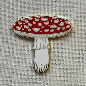 May include: A red and white enamel pin shaped like a mushroom with white spots. The mushroom has a gold outline.