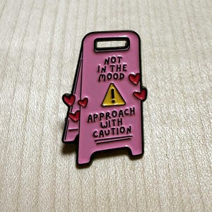 May include: A pink enamel pin shaped like a caution floor sign. The sign features a yellow triangle with an exclamation point and the black text: NOT IN THE MOOD, APPROACH WITH CAUTION. Several small red hearts are attached to the sides of the sign.
