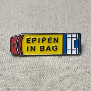 K&ouml;nnte beinhalten: Eine gelbe und blaue Emaille-Anstecknadel in Form eines Epipens mit dem Text "EPIPEN IN BAG" in schwarzen Buchstaben.