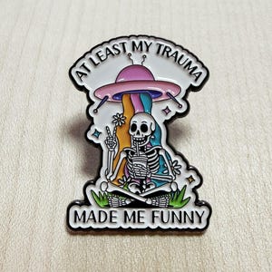 Könnte beinhalten: Ein Metall-Emaille-Pin mit einem weißen Skelett, das im Schneidersitz sitzt, ein Getränk hält und ein Friedenszeichen macht. Über dem Skelett schwebt ein rosa UFO mit einem Regenbogenstrahl. Der Text lautet AT LEAST MY TRAUMA MADE ME FUNNY.