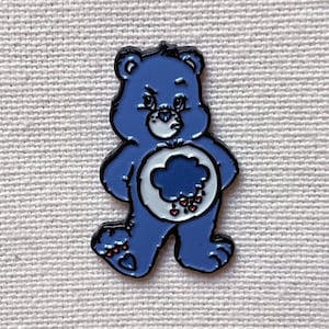 May include: A blue Care Bear enamel pin with a grumpy expression and a dark blue cloud with raindrops and hearts on its belly.
