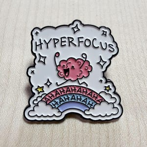 Puede incluir: Un pin de esmalte blanco con forma de nube y contornos negros. Presenta un cerebro rosa de dibujos animados con los brazos levantados, el texto "HYPERFOCUS" en negro y un arcoíris colorido debajo con el texto "HAHAHAHA" repetido en negro.