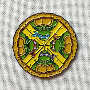 Può includere: Uno spillo in smalto giallo e verde con quattro Tartarughe Ninja Adolescenti in un cerchio. Le tartarughe sono rivolte verso l'esterno con la bocca aperta e gli occhi chiusi. Lo spillo è circondato da un bordo rosso e dorato.