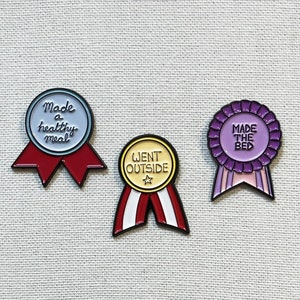 May include: Three enamel pins with colourful ribbons. The pins feature the text "Made a healthy meal", "Went Outside", and "Made the Bed".