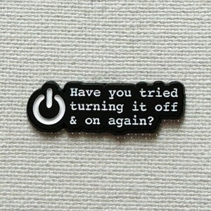 Op de afbeelding: Zwart-witte emaille pin met een aan/uit-knopicoon en de tekst "Heb je geprobeerd om het uit en weer aan te zetten?"