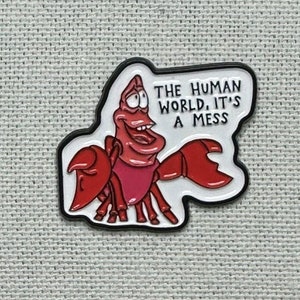 Pode incluir: Um broche de esmalte vermelho e branco com um caranguejo de desenho animado com o texto "O mundo humano, é uma bagunça".