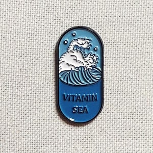 May include: A blue and white enamel pin shaped like a pill with a wave and the text "Vitamin Sea" on it.