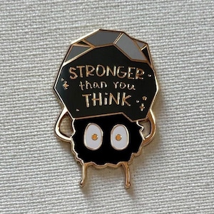 May include: Black and gray enamel pin with a black and white cartoon character. The character has a gray rock on its head with the text "STRONGER than you THINK" in white.