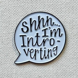 May include: White enamel pin with a speech bubble design. The text inside the bubble reads "Shhh... I'm Intro-verting".