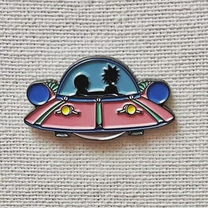 May include: A pink and blue enamel pin in the shape of a flying saucer. The pin features a blue dome with black silhouettes of two characters. The saucer has blue lights and yellow accents.