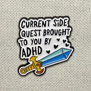 Pode incluir: Um broche de esmalte branco com texto preto que diz CURRENT SIDE QUEST BROUGHT TO YOU BY ADHD, decorado com pequenos corações pretos e um gráfico de uma espada com punho dourado e lâmina azul clara, com contorno em metal preto.