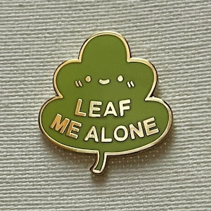 May include: Green leaf-shaped enamel pin with a gold outline. The pin has a smiling face and the text "LEAF ME ALONE" in gold lettering.
