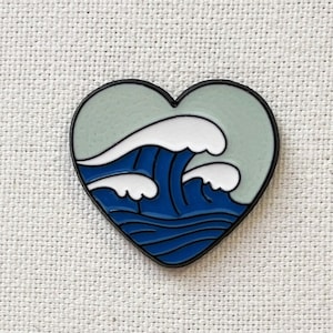 Könnte beinhalten: Eine blaue und weiße Emaille-Anstecknadel in Herzform mit einem Wellendesign im Inneren. Das Herz ist hellblau und die Welle ist dunkelblau mit weißem Schaum.