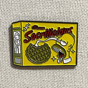 May include: A yellow enamel pin featuring a cartoon character with a yellow face and blue hair eating a green waffle. The text "Mmm Sacrillicious" is written in red and white on the box. The pin is a parody of a popular breakfast cereal.