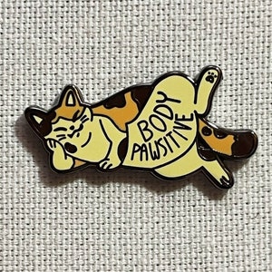 May include: A yellow and brown cat with a black nose and eyes is lying on its back with its paws in the air. The cat has a speech bubble that says "Body Pawsitive".