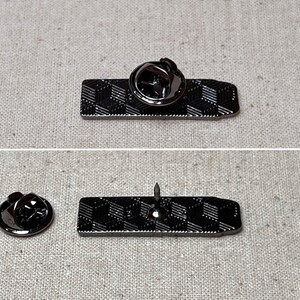 K&ouml;nnte beinhalten: Eine schwarze Metallstifte mit einem geometrischen Muster. Der Stift hat einen Sicherheitsverschluss auf der R&uuml;ckseite.