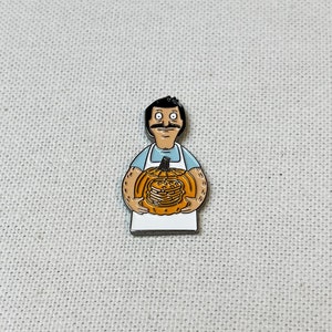 K&ouml;nnte beinhalten: Eine Emaille-Anstecknadel mit einem Cartoon-Mann mit Schnurrbart, der einen K&uuml;rbis h&auml;lt. Der K&uuml;rbis ist orange und hat ein geschnitztes Gesicht. Der Mann tr&auml;gt ein blaues Hemd und eine wei&szlig;e Sch&uuml;rze.